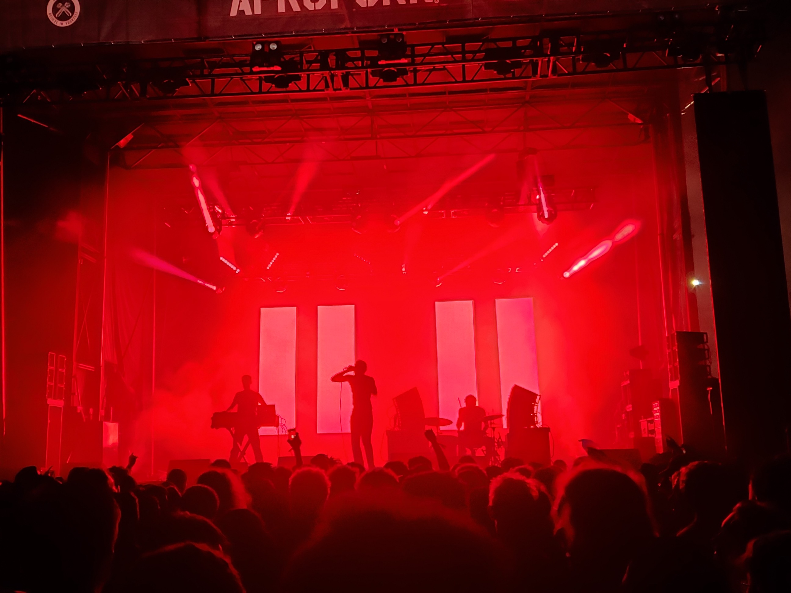 a hazy stage lit with red lights, silhouette of three performers, the backs of the audiences heads