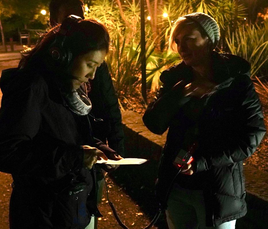 the artist explaining the installation to a listener wearing headphones connected to a small radio and looking at a paper program