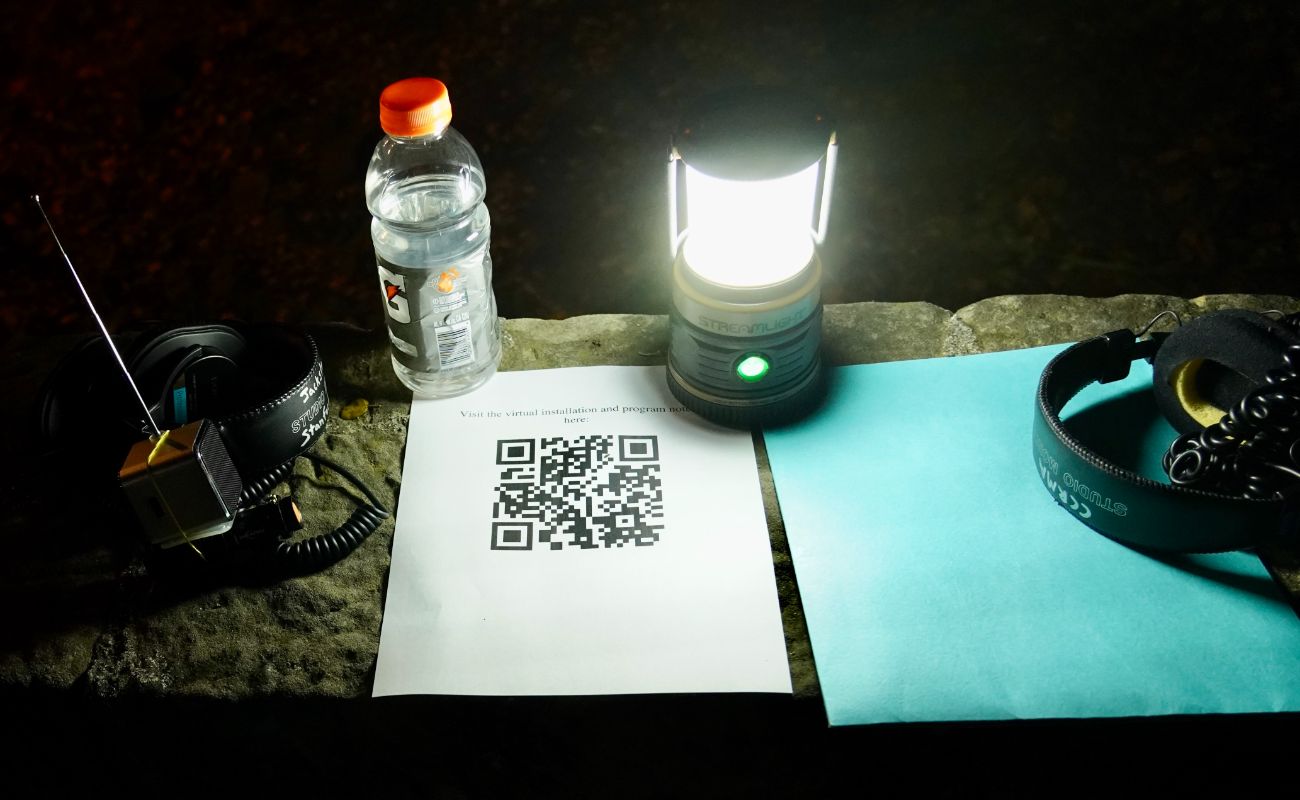 stone ledge with electric lantern lighting up a blue folder, headphones connected to radio receivers attached to flashlights, and paper with a QR code and the text Visit the virtual installation and program notes here