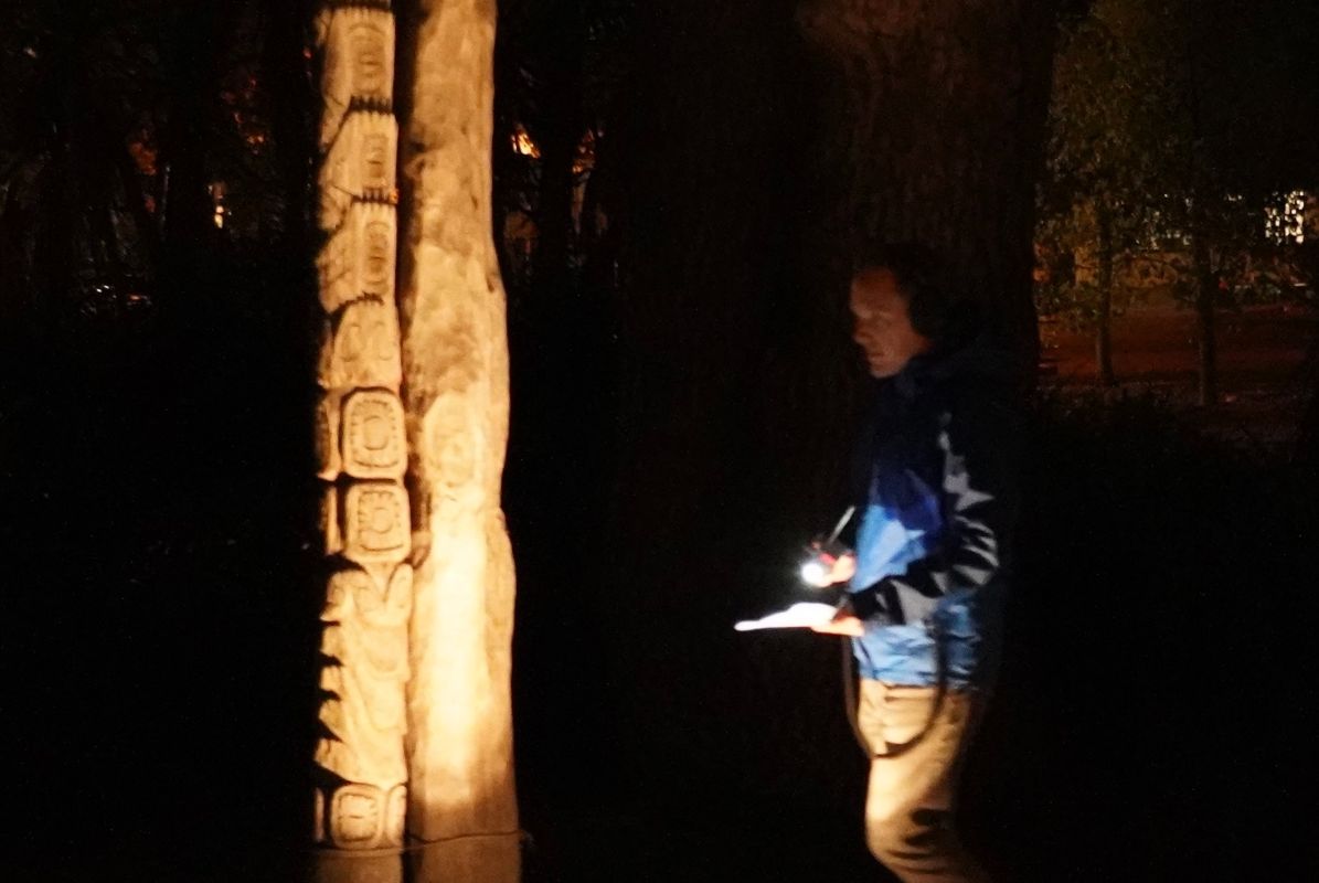 listener approaching a tall wooden sculpture, wearing headphones and pointing his flashlight at a program, dark trees and the nearby street in the background