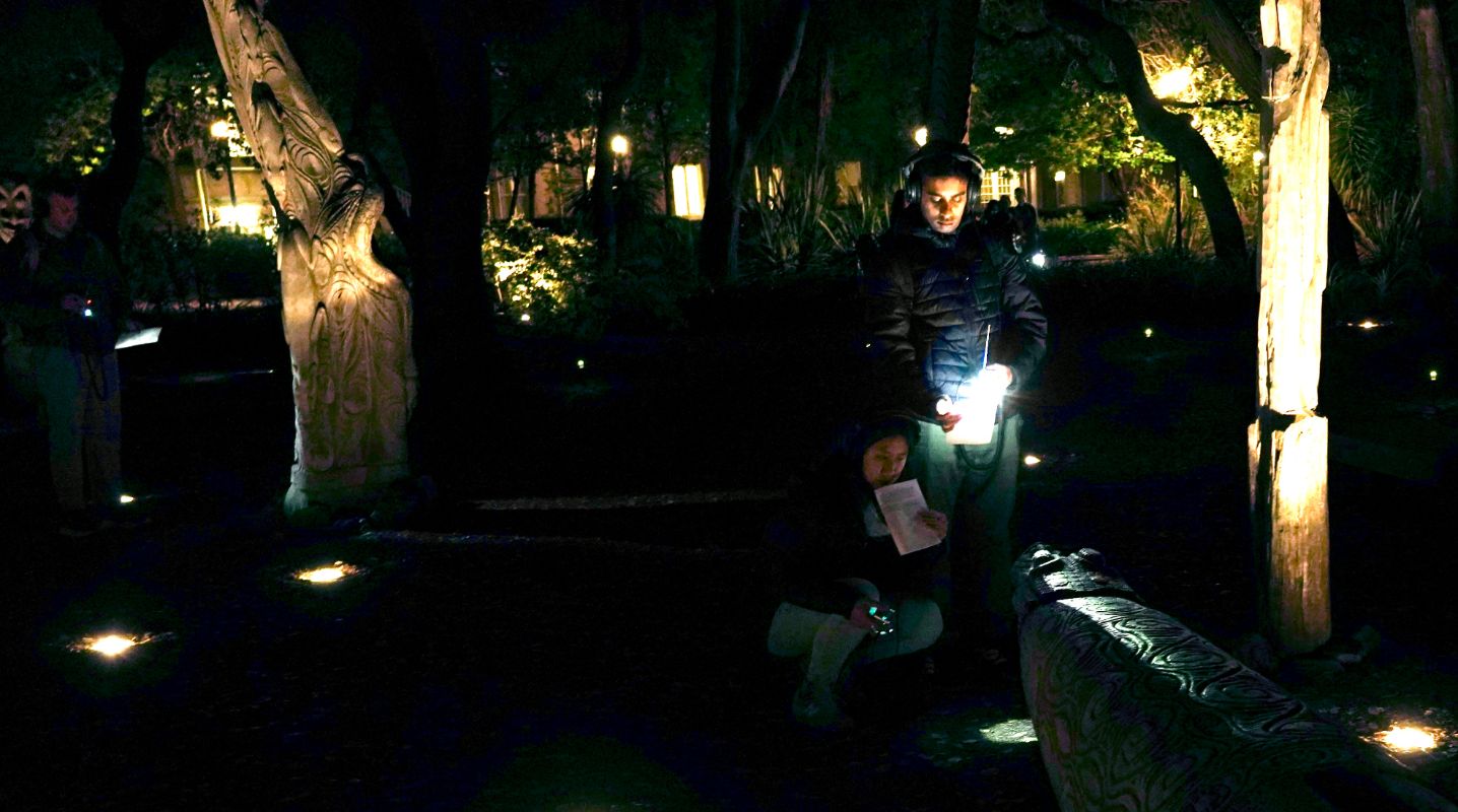 two listeners near a crocodile-shaped bench, both with headphones on, one squatting with a radio receiver and program in her hands, the other standing and shining his flashlight towards the crocodile's head