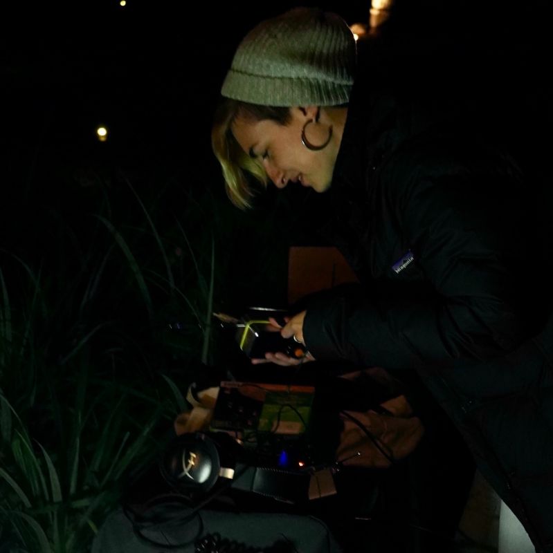 the artist leaning over a spread of various radios, flashlights, and headphones, surrounded by plants